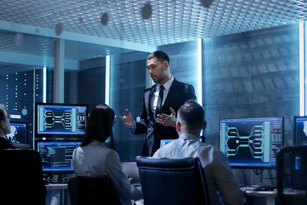 Supervisor Holds Briefing for His Employees in System Control Center Full of Monitors and Servers. Possibly Government Agency Conducts Investigation.