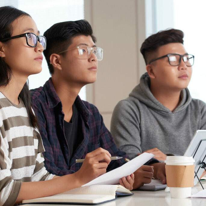 Profile view of concentrated Asian students sitting at spacious classroom with panoramic windows and looking at blackboard